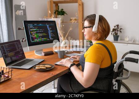 Young pretty female software developer in wheelchair sitting by desk in front of laptop and computer monitor while working with data Stock Photo