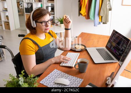 Young female programmer sitting by desk at home, eating cookies with tea and working with data in front of computer screen and laptop Stock Photo