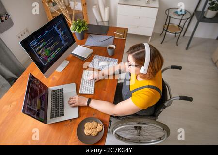 Young female programmer in wheelchair looking at data on laptop display while sitting by desk in home environment and developing software Stock Photo