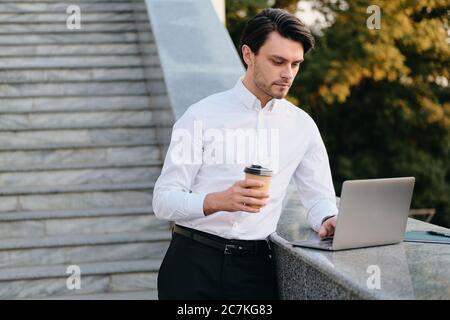Young beautiful serious bearded brunette man in white shirt holding cup of coffee to go in hand thoughtfully working on laptop in city park Stock Photo