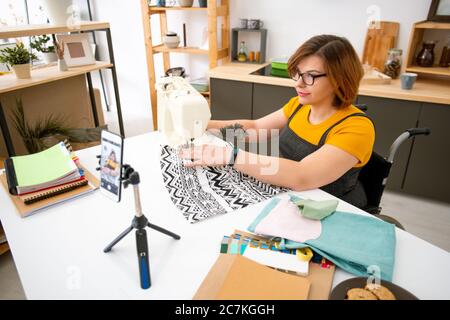 Young disable female in wheelchair sitting by sewing machine at home while making livestream masterclass for online audience Stock Photo
