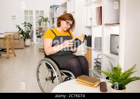 Young disable female in casualwear and eyeglasses sitting on wheelchair by shelf and reading book while getting home education Stock Photo