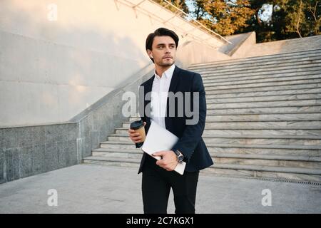 Young handsome bearded brunette man in white shirt and classic suit with wireless earphones holding cup of coffee to go and laptop in hands Stock Photo