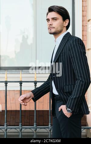 Young handsome bearded brunette man in white shirt and classic striped jacket dreamily looking aside standing on city street Stock Photo