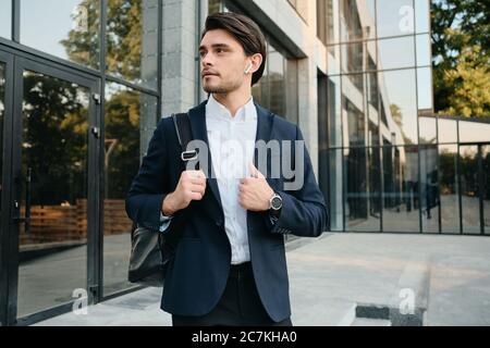 Young handsome bearded brunette man in white shirt and classic suit with wireless earphones and backpack on shoulder dreamily looking aside with Stock Photo