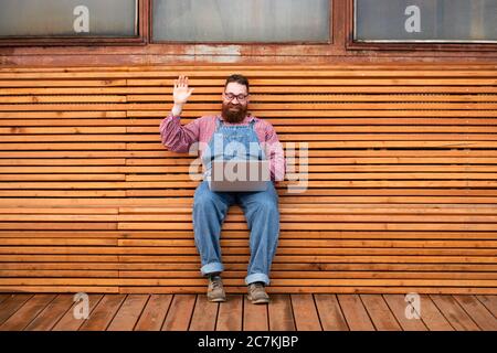 Portrait of cheerful brutal bearded hipster man in blue overalls, checked shirt using on laptop talks with his girlfriend and waving his hand
