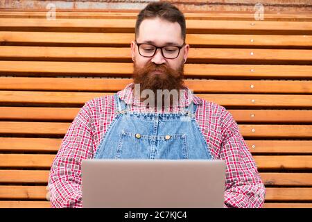 Close up portrait of serious brutal bearded hipster man in blue overalls, checked shirt working on laptop sitting on bench outdoors. Distance job Stock Photo