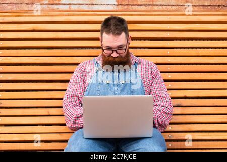Close up portrait of serious brutal bearded hipster man in blue overalls, checked shirt working on laptop sitting on bench outdoors. Distance job Stock Photo