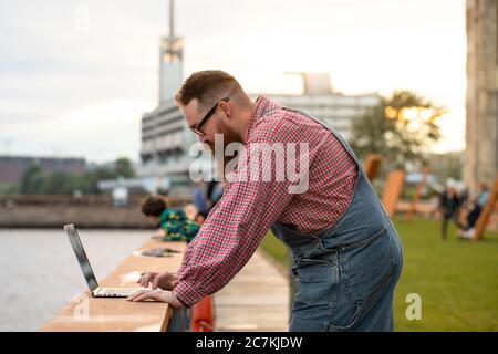 Portrait of brutal bearded man freelancer wearing blue overalls and checked shirt working on laptop at embankment outdoors. Distance job, remote work. Stock Photo