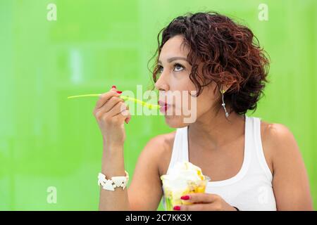 Close-up of a brown-skinned girl eating an ice cream on a green background. Space for text. Selective focus. Summer and healthy lifestyle concept. Stock Photo