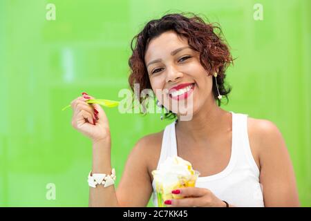 Close-up of smiling latin woman holding an ice cream on a green background. Space for text. Selective focus. Summer and lifestyle concept. Stock Photo