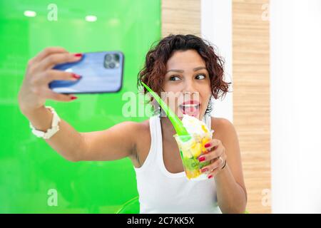 Portrait of a beautiful, brown-skinned woman taking a selfie with her smartphone while holding an ice-cream cup in her hand. Selective focus. Summer a Stock Photo