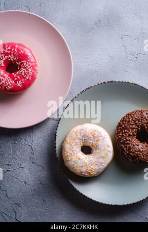 Three donuts on plates, chocolate, pink and vanilla donut with sprinkles, sweet glazed dessert food on concrete textured background, angle view