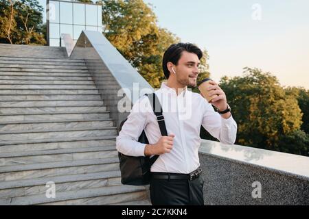 Young smiling bearded brunette man in white shirt with wireless earphones standing with cup of coffee to go in hand and backpack on shoulder happily Stock Photo