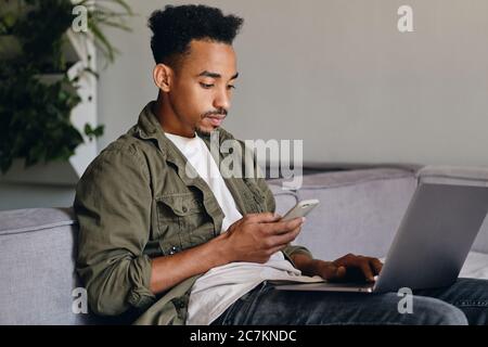 Young serious African American guy with laptop thoughtfully using cellphone in co-working space Stock Photo
