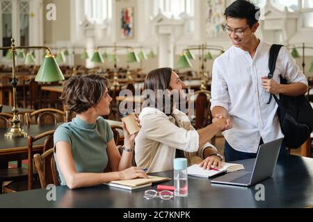 Group of young casual students happily greeting each other while studying in library of university Stock Photo