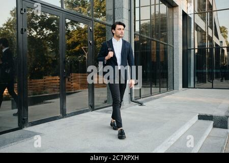 Young attractive bearded brunette man in white shirt and classic suit with wireless earphones and backpack on shoulder thoughtfully looking aside Stock Photo
