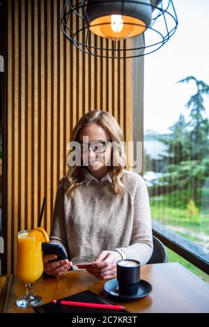Elegant blonde woman sitting at cafe holding mobile phone and credit card signing up on website. Businesswoman paying with credit card while shopping Stock Photo