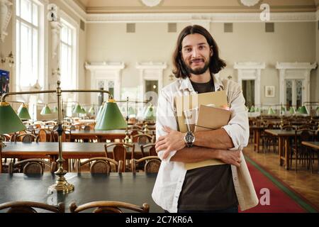 Young casual male student with notepads happily looking in camera in library of university Stock Photo
