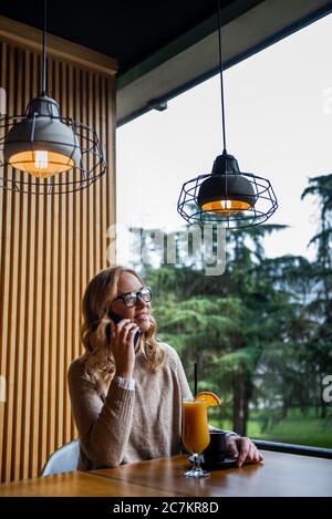 Young charming woman calling with cell telephone while sitting alone in coffee shop during free time, attractive female with cute smile having talking Stock Photo