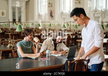 Young asian student in eyeglasses with backpack coming to study with friends in library of university Stock Photo
