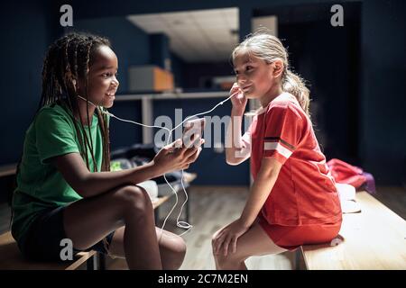 Two smiling girl soccer player before training listen music on phone in locker room. Stock Photo