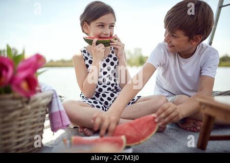 Cute kids  eating fresh watermelon on the pier.  Girl and boy enjoying together at lake. Stock Photo