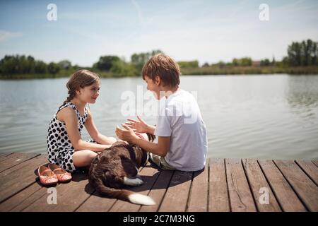 Happy childhood in nature.Little children with dog sitting together on wooden near pond and talking. Stock Photo