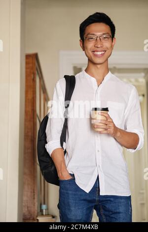 Young attractive asian male student in eyeglasses with backpack and coffee to go joyfully looking in camera in university Stock Photo