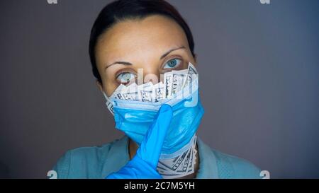 Woman doctor or nurse covering her mouth with fifty dollar bills sticking out of her medical face mask with a rubber gloves hand gestures silence Stock Photo