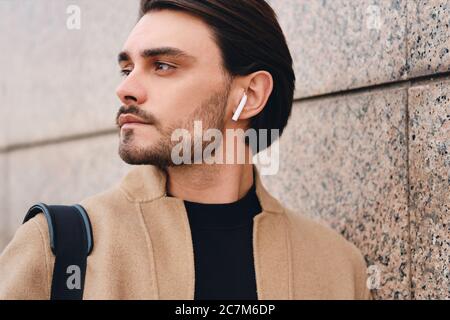 Side view of young man in coat with wireless earphones wistfully looking away outdoor Stock Photo