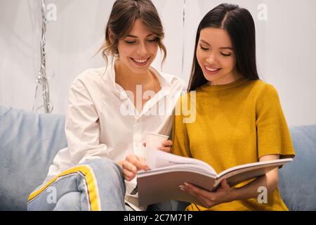 Two beautiful stylish girls happily reading fashion magazine on sofa in showroom Stock Photo