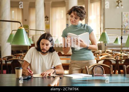 Attractive female tutor with book explaining to young student new theme during lesson in library of university Stock Photo