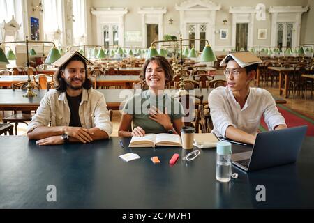 Group of young cheerful multinational students joyfully fooling around with book on head while studying together in library of university Stock Photo