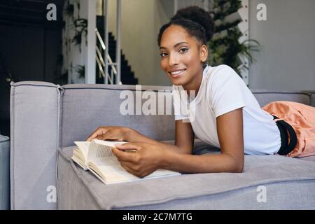 Pretty smiling African American girl lying on sofa with book joyfully looking in camera at home Stock Photo