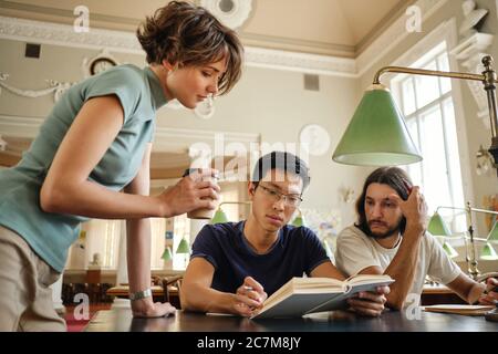 Young attractive tutor helping multinational students with study during lesson in library of university Stock Photo