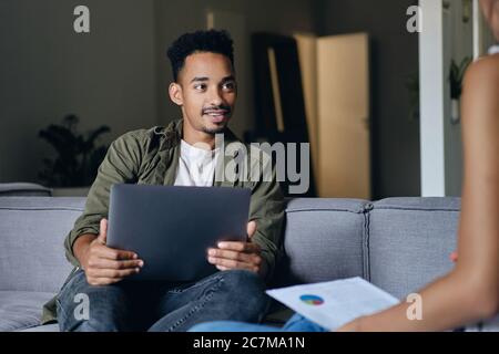 Young attractive casual African American man with laptop dreamily working with colleague in co-working space Stock Photo