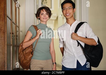 Two young attractive international students with backpacks happily looking in camera while standing in corridor of university Stock Photo