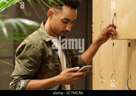 Side view of young attractive African American guy dreamily using cellphone in locker room Stock Photo