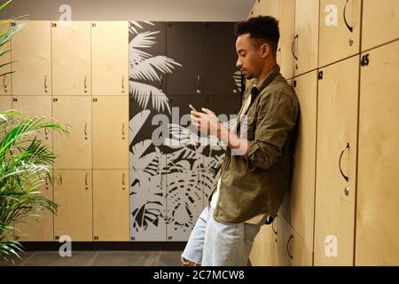 Young serious casual African American guy thoughtfully using cellphone in locker room alone Stock Photo