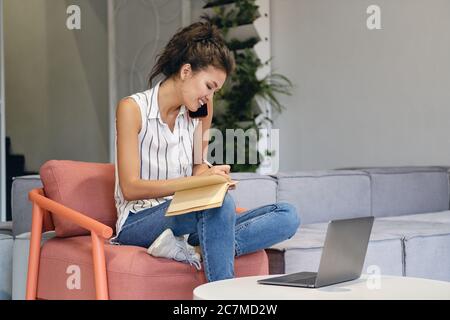 Pretty smiling casual asian girl talking on cellphone writing notes during work with laptop in co-working space Stock Photo