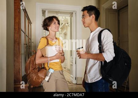 Two young attractive casual international students with backpacks and notepad happily talking in corridor of university Stock Photo