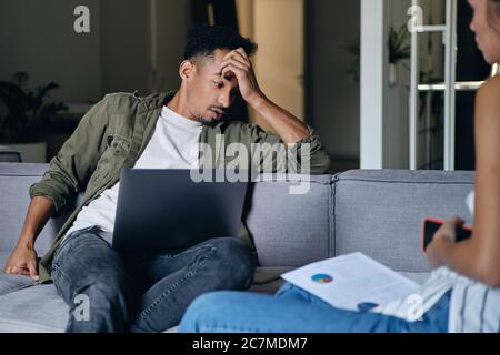 Young casual African American man with laptop tiredly working with colleague in co-working space Stock Photo
