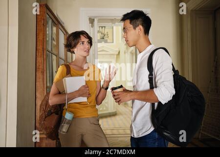 Two young international students with backpacks and notepad thoughtfully discussing study in corridor of university Stock Photo