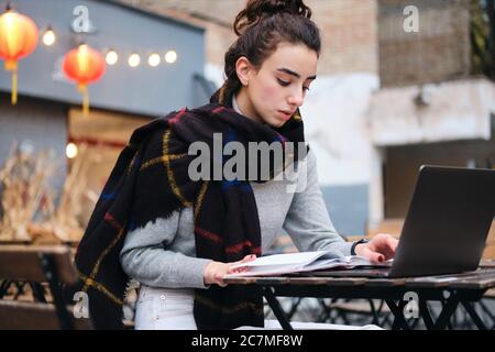 Serious brunette girl with scarf thoughtfully reading book studying with laptop in cafe on city street Stock Photo