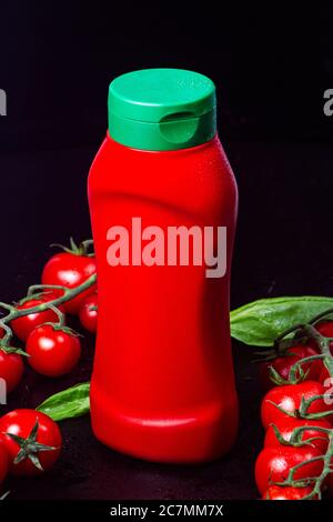 bottle, tube of red ketchup with a green cap and two ripe branches of cherry tomatoes with spinach with drops of dew water on a black embossed table. Stock Photo