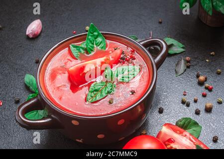 Tomato soup in a small deep plate with spices, basil and tomato on a black table. copy space, close up, side view Stock Photo