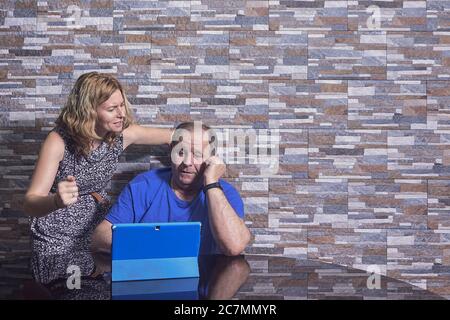 Aged man with a blue tablet in front of him talking on the phone and his wife disturbing him Stock Photo