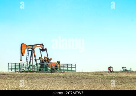 Close--up of oil well pump jack out in a field enclosed in a metal cattle fence with two other pumping wells in the distance all set on the horizon of Stock Photo