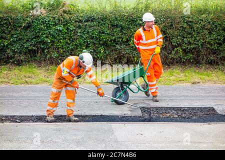 Workmen and woman doing road repairs repairing damaged uneven road surface and pot holes with tarmac in a Cheshire country lane road England UK Stock Photo